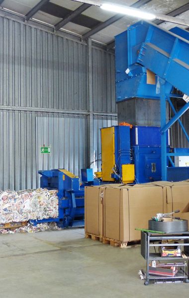 Innenraum einer Halle von Xpress Paper GmbH, Paper Recycling Management. Paletten mit Altpapier stehen zur Verarbeitung bereit. Förderband bewegt das Altpapier zur Presse. Diese presst den Rohstoff zu Ballen zusammen und bindet diese für Transport.