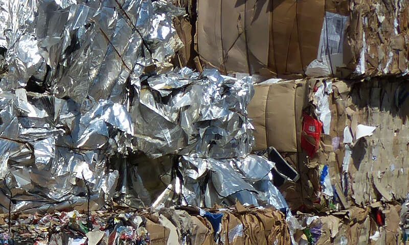 Glänzende silberne Kunststofffolie aus Gewerbebetrieb zum Rohstoffballen gepresst, mit Draht gebunden und zu einem futuristisch wirkenden Berg gestapelt. Daneben stehen Sekundärrohstoffballen aus gepressten Karton aus Verpackungen im Einzelhandel.