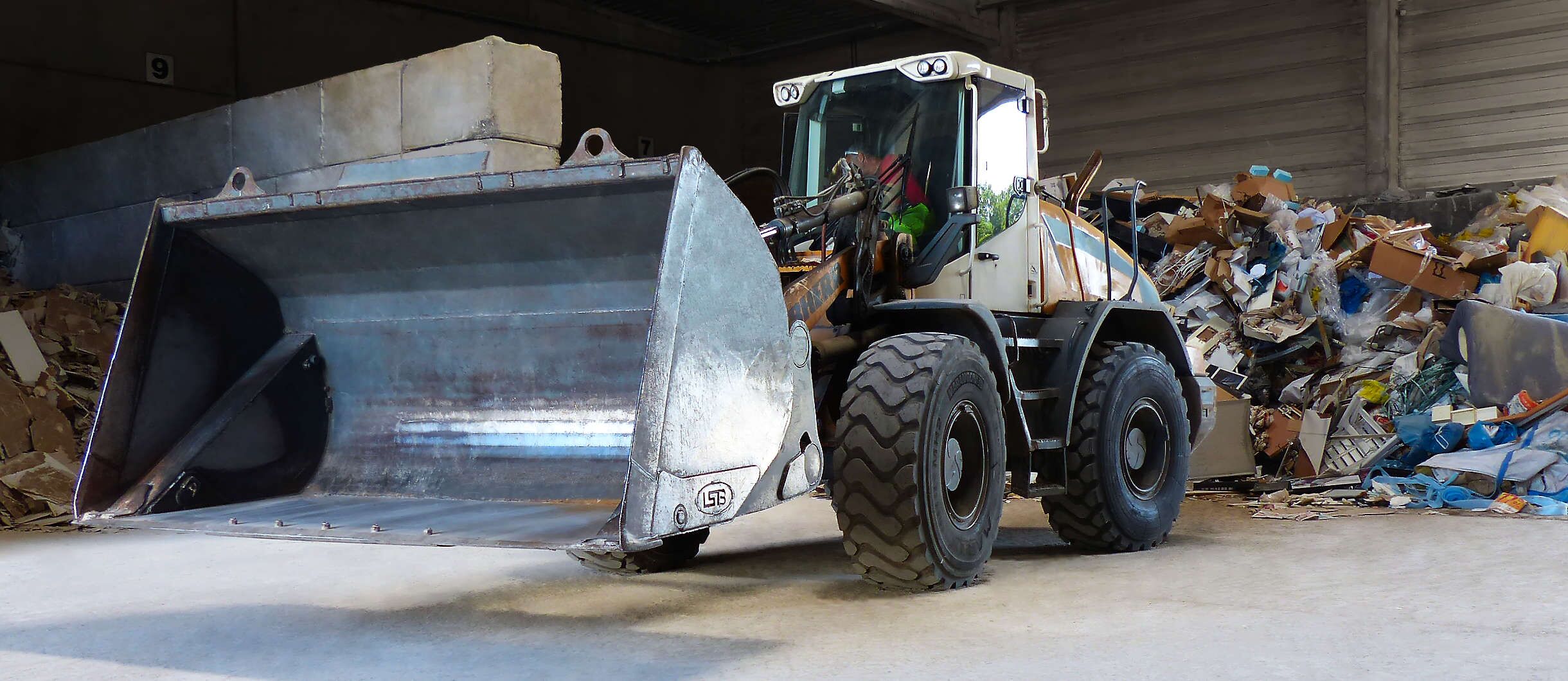 In der Betriebshalle für Altpapier- und Plastik-Sortierung steht ein mächtiger Radlader mit einer riesigen angehobenen Mulde. Im Hintergrund liegt zu unterschiedlichen Haufen sortierter Abfall für die Rohstoffwiedergewinnung. Er wird gepresst & recycelt.