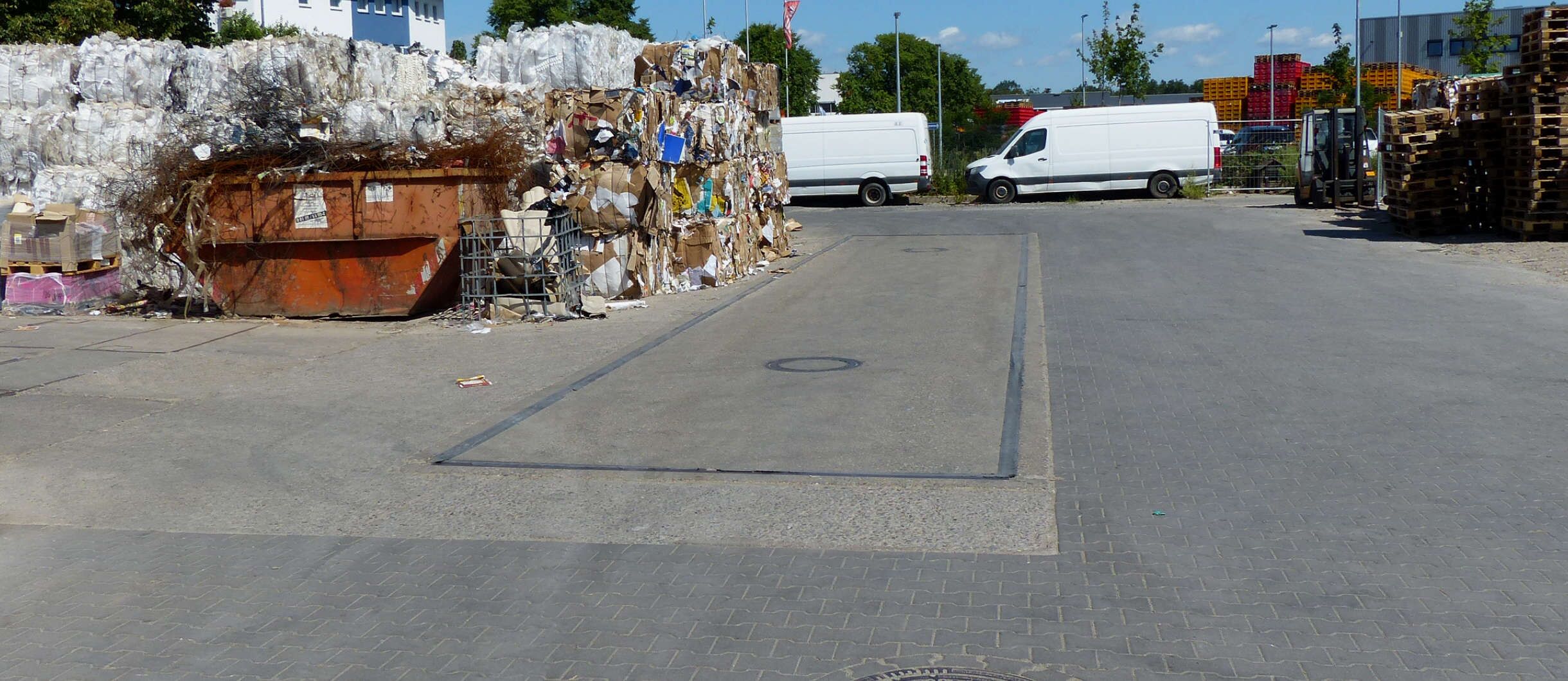 Auf dem Gelände des Altpapier-Recyclingunternehmens Xpress Paper stehen zwei weiße Sprinter, Paletten stehen gestapelt. Eine LKW-Waage ist in den Boden einbetoniert. Daneben stehen Container und aus Papier, Karton und Kunststoffen gepresste Ballen.