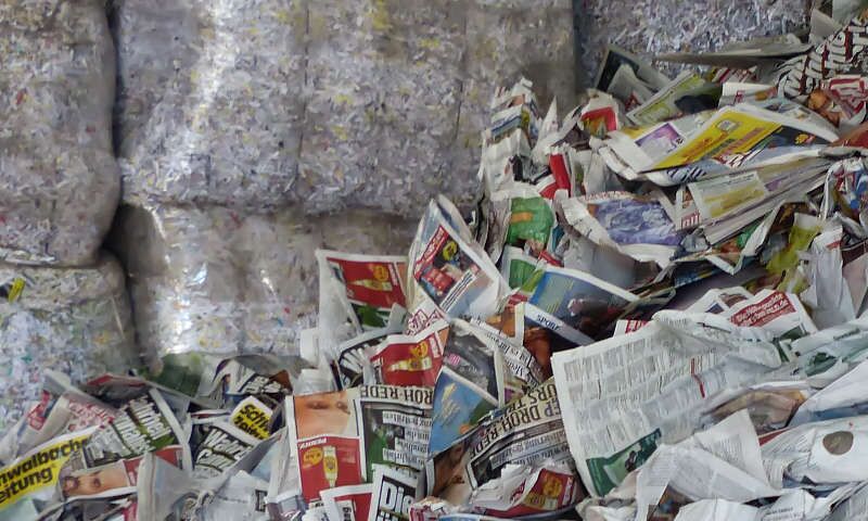 Im Vordergrund liegt unsortiertes Zeitungspapier. Im Hintergrund sind Pellets in Ballen gebunden auf Paletten gestapelt. Abfall wurde sortiert, verwertet, gepresst, verpackt. Ersatzbrennstoffe (EBS) bzw. Sekundärbrennstoffe (SBS) für den Verkauf bereit.