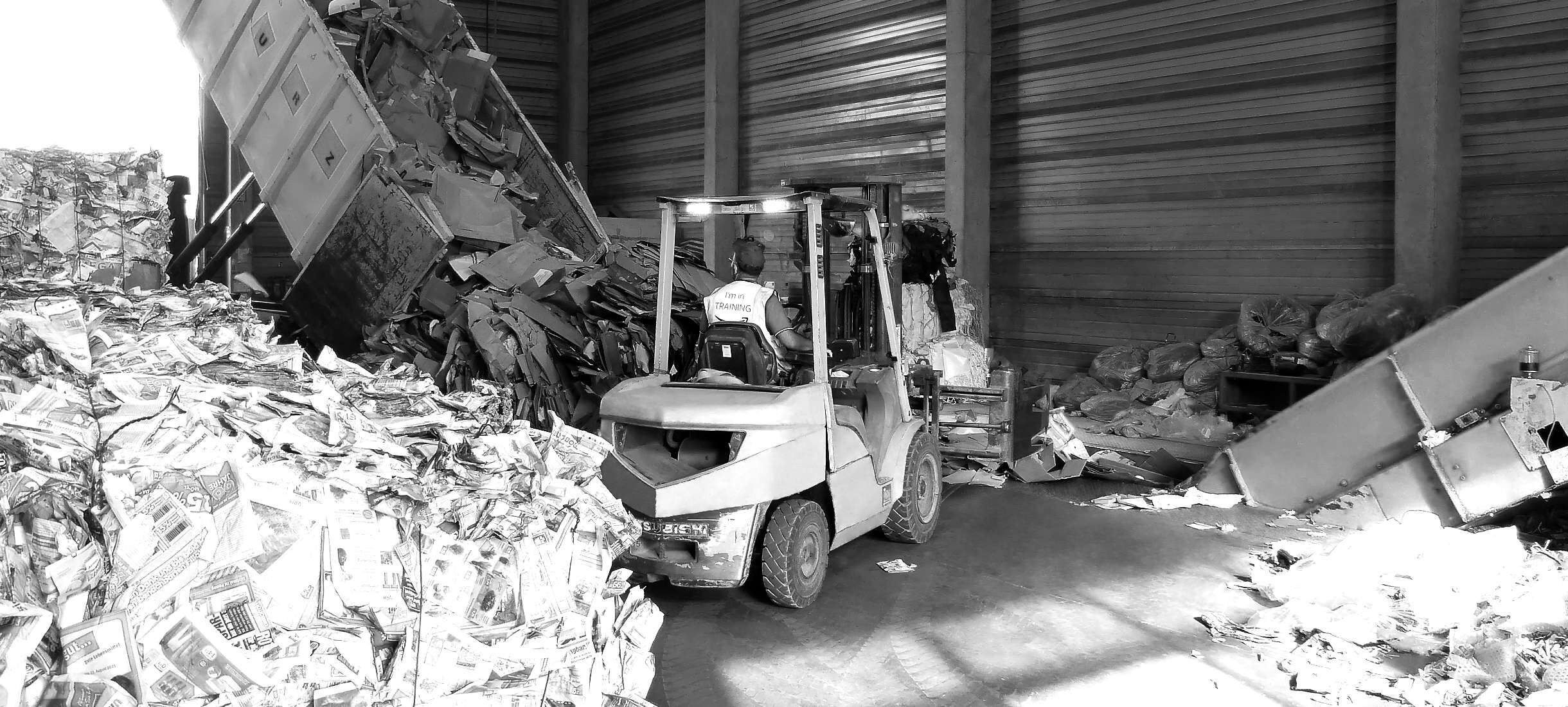 Ein LKW entlädt große Mengen an Kartons aus Wellpappe in der Recyclinghalle eines Entsorgungsunternehmens. Ein Mitarbeiter auf Vollzeit fährt mit einem Gabelstapler zum Haufen aus Pappe. Die Halle ist voll mit Altpapier und Plastikverpackungen.