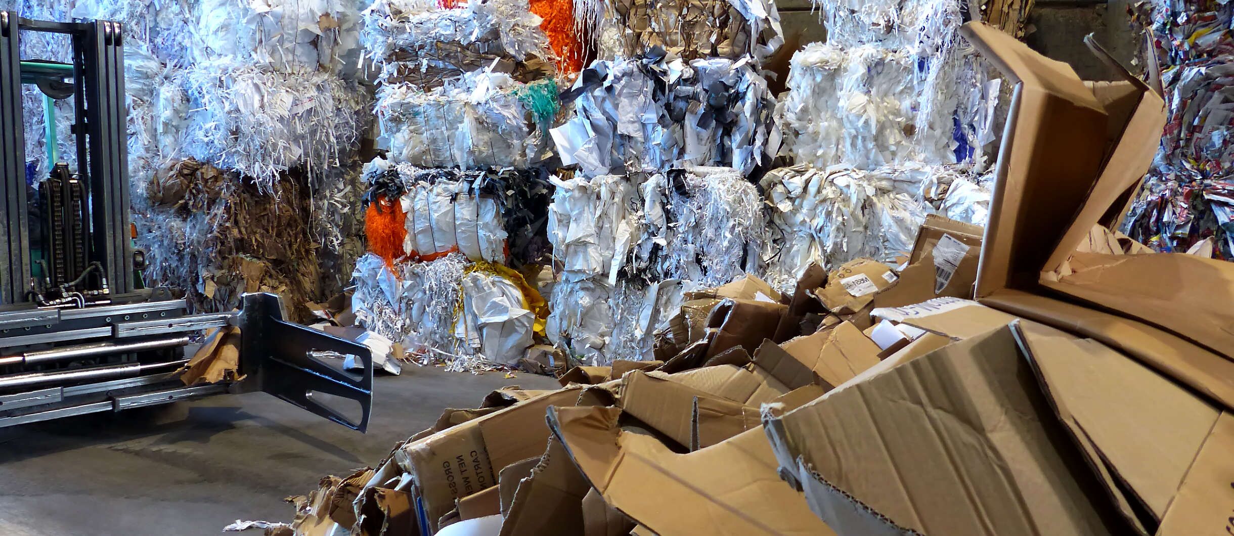 In der Halle mit der Papierpresse stehen Papierballen aus Druckerei-Resten und gepressten Zeitungen aus Verlagsrückständen. Gabelstaplerfahrer trägt einen Berg aus Verpackungen von Supermärkten und Pappkartons aus Wellpappenwerken zu der Ballenpresse.