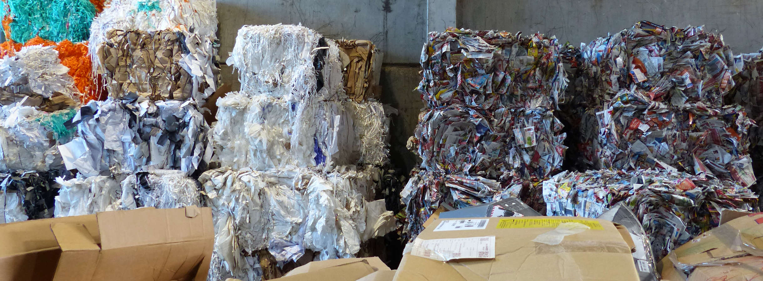 In der Produktionshalle des Entsorgungsfachbetriebs für Frankfurt-Rhein-Main stehen bunte Altpapier-Ballen aus Zeitungs- oder Druckereiabfällen und Karton-Ballen an der Wand gestapelt. Im Vordergrund liegen Wellpappenreste zur Weiterverarbeitung bereit.