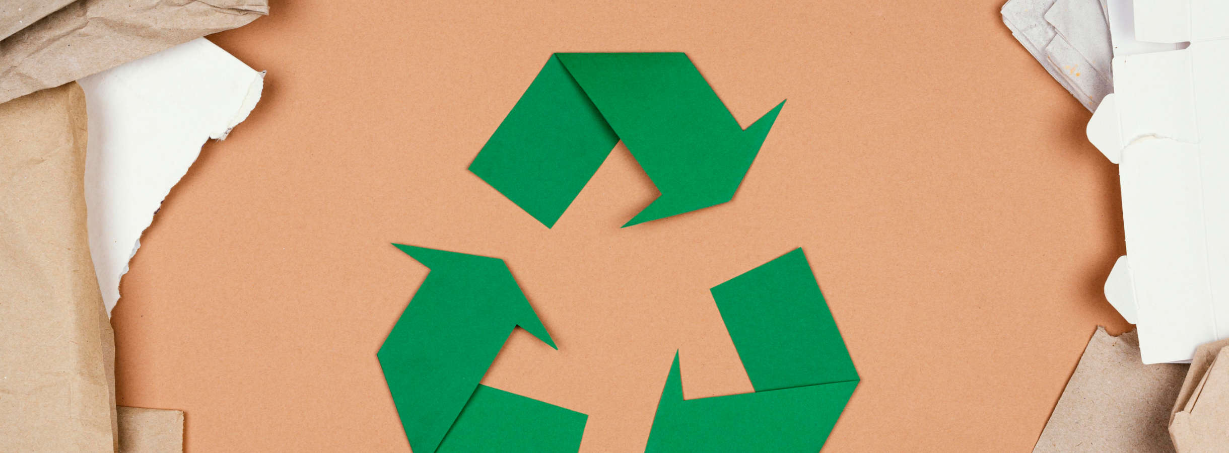 Recyclingsymbol mit drei Pfeilen im Kreis für umweltfreundliche Altpapierverwertung und Pappeverwertung. Das grüne Zeichen wurde aus Papier ausgeschnitten, angeordnet und liegt umgeben von Papierresten auf ausgebreiteter Pappe.