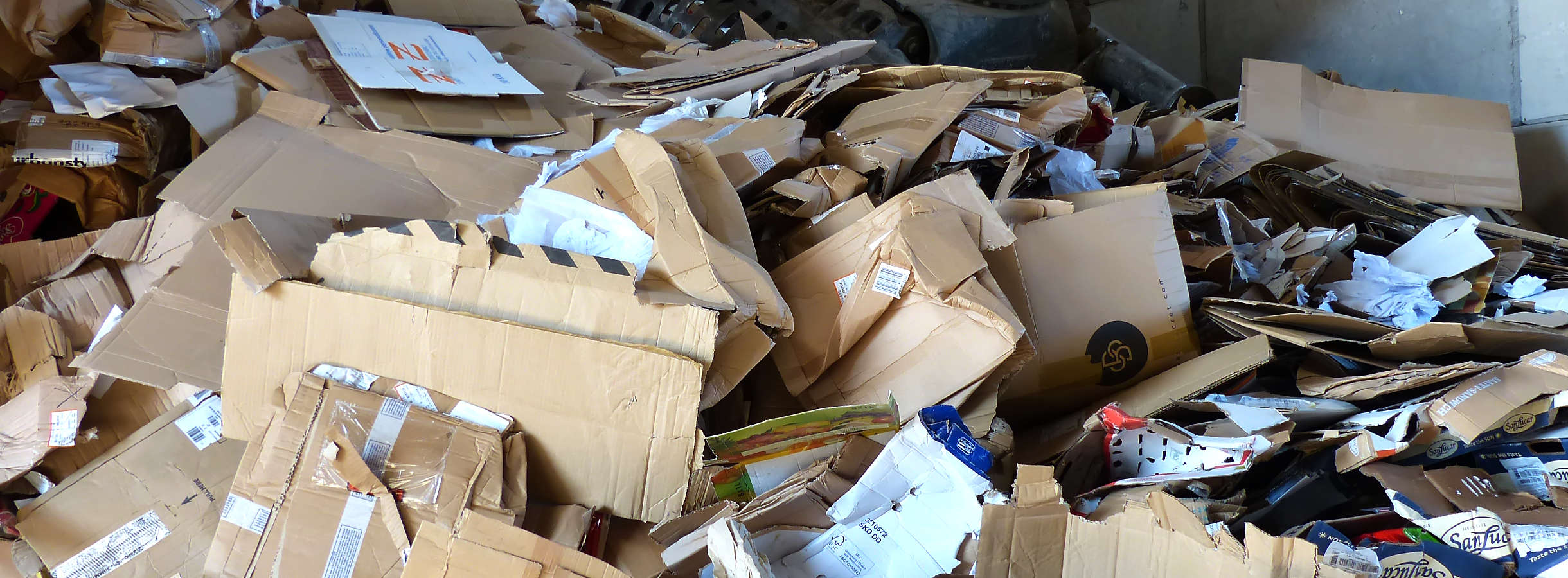 Entsorgte beige und bunte Wellpappe zu einem riesigen Berg aufgehäuft. Darauf liegt der Greifarm eines Baggers. Es sind kaputte Pappkartons, Wellpappenreste oder Wellpappenfehlproduktionen gesammelt und entsorgt in Wellpappenwerken.