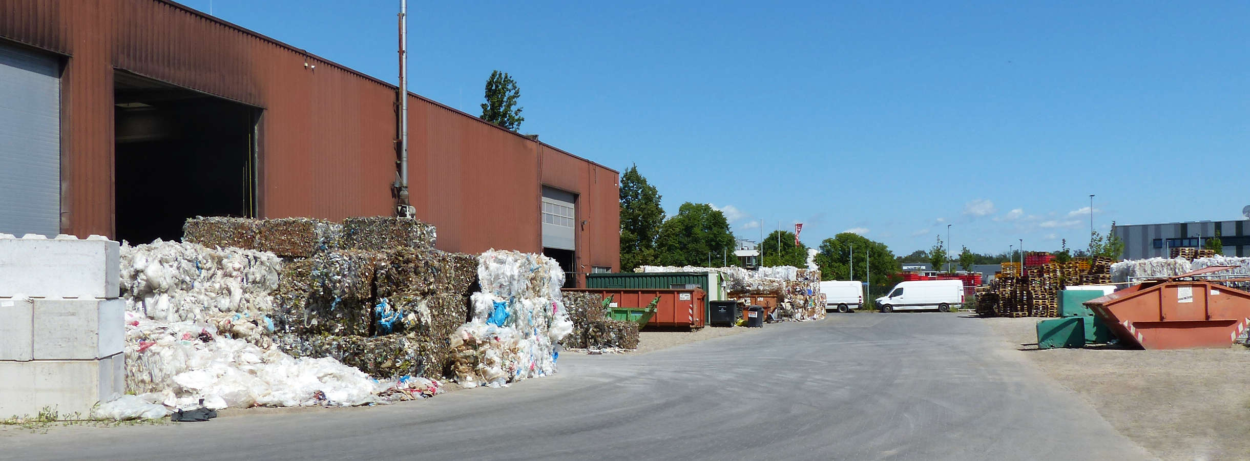 Recyclinghalle der Xpress Paper GmbH für Papier-, Plastik-, Papperecycling in Terrakotta. Drei Tore führen in das Innere. Auf dem Gelände stehen bunte Abfall-Container verschiedener Anforderungen, Paletten, mehrere Ballen gepresster Recycling-Rohstoffe.