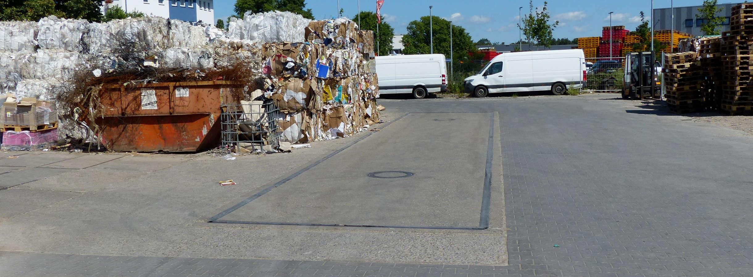 Fahrzeugwaage auf dem Gelände des Altpapier-Recyclingunternehmens Xpress Paper. Darauf werden große & kleine LKWs mit angelieferten sekundären Papier- oder Plastikrohstoffen vor dem Handel gewogen. Daneben stehen Paletten, Container und Altpapierballen.