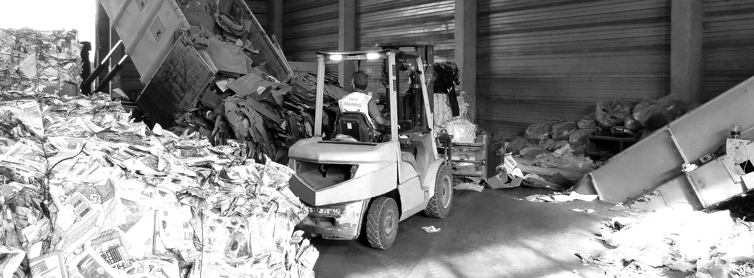 Mitarbeiter der Firma fährt mit einem Gabelstapler zwischen riesigen Haufen aus Altpapier, Pappe und Plastik-Verpackung. Er fängt mit der Gabel die kürzlich ausgeschüttete Pappe und transportiert den Rohstoff zum Recyceln in eine Papier-Presse.