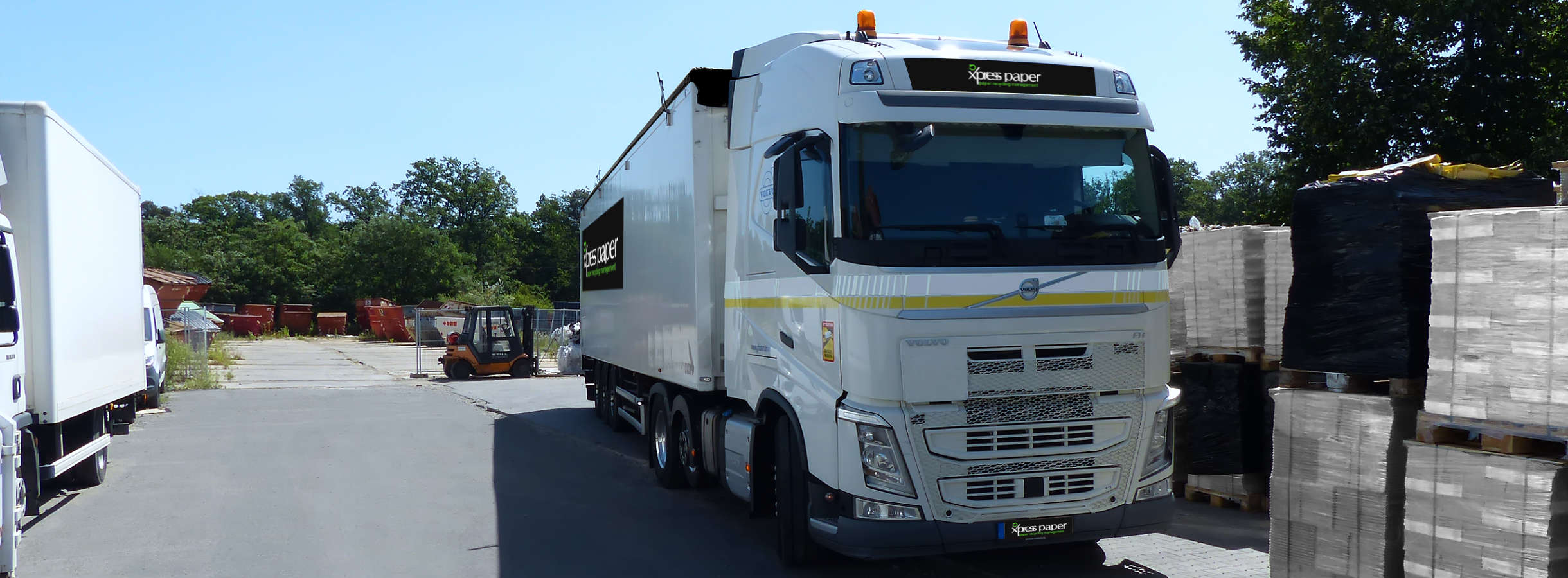 LKW parkt auf dem Gelände des Entsorgungsbetriebs Xpress Paper GmbH. Er hat Altpapier zur Weiterverarbeitung angeliefert. Anschließend wurde er mit gepressten Papierballen beladen. Hinterm LKW steht ein Gabelstapler, neben viele Paletten mit Altpapier.