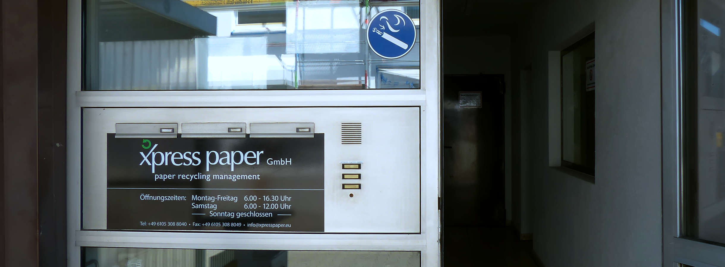 Eingang ins Bürogebäude des Recyclingunternehmens Xpress Paper GmbH in Mörfelden-Walldorf. Links neben der offenen Eingangstür sind Klingeln und drei Postkästen angebracht. Darunter befindet sich das Firmenlogo mit Kontaktdaten und Öffnungszeiten.