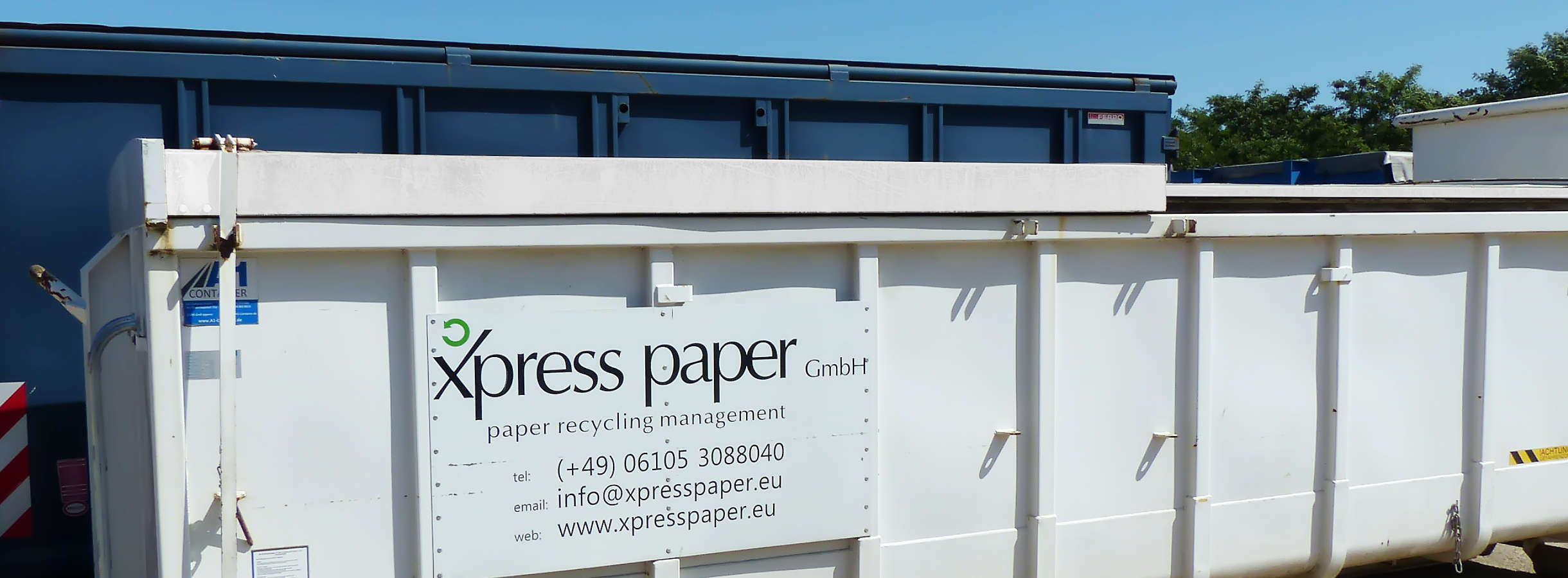 Große weiße und blaue Altpapier-Container vom Entsorgung- und Recyclingbetrieb für Altpapier in Mörfelden-Walldorf. Auf einem weißen Container ist das Logo von Xpress Paper mit Kontaktdaten abgebildet. Container für Elektroschrott stehen weiter hinten.