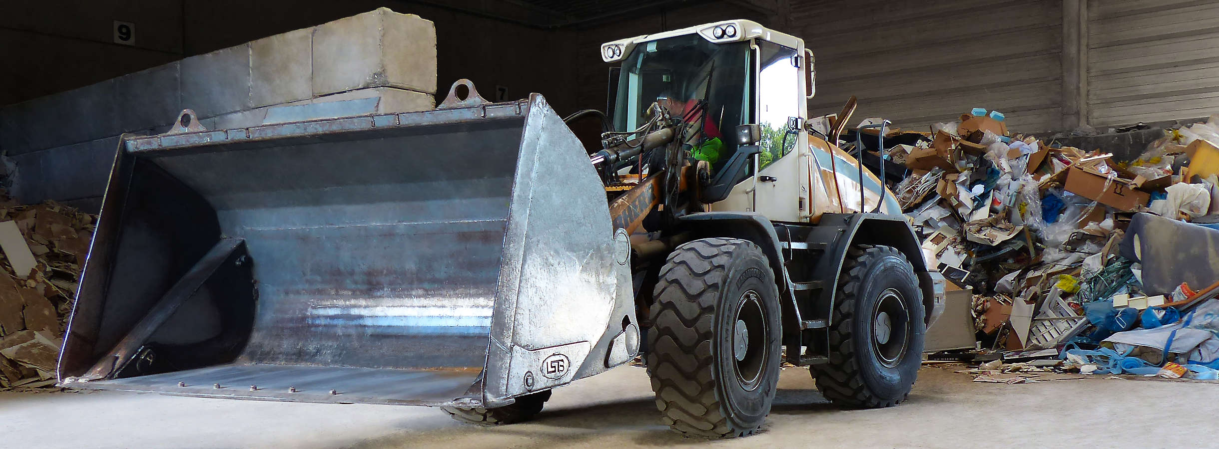 Großer Bagger mit einem riesigen Radlader steht mitten in der Lagerhalle vom Xpress Paper Altpapier-Recyclingunternehmen. Der Baggerfahrer sortiert angelieferten Karton- und Pappe-Abfall in zugewiesene Ecken der Halle zur Vorbereitung vom Recycling.