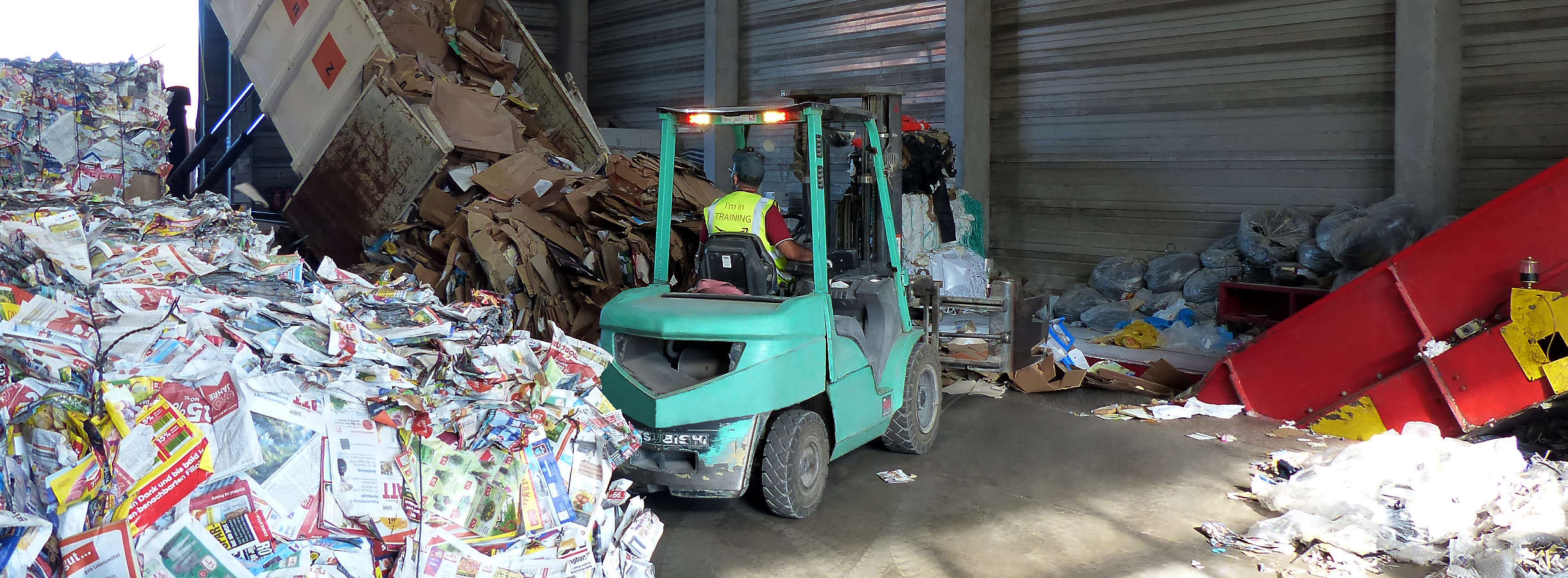 In einer Produktionshalle des Recyclingunternehmens im Großraum Frankfurt wird Altpapier und Pappe zu Ballen gepresst, gebunden. Ein weißer LKW lädt Kartonage ab. Mitarbeiter aufm Gabelstapler sortiert die Rohstoffe und gibt sie in eine Papier-Presse.