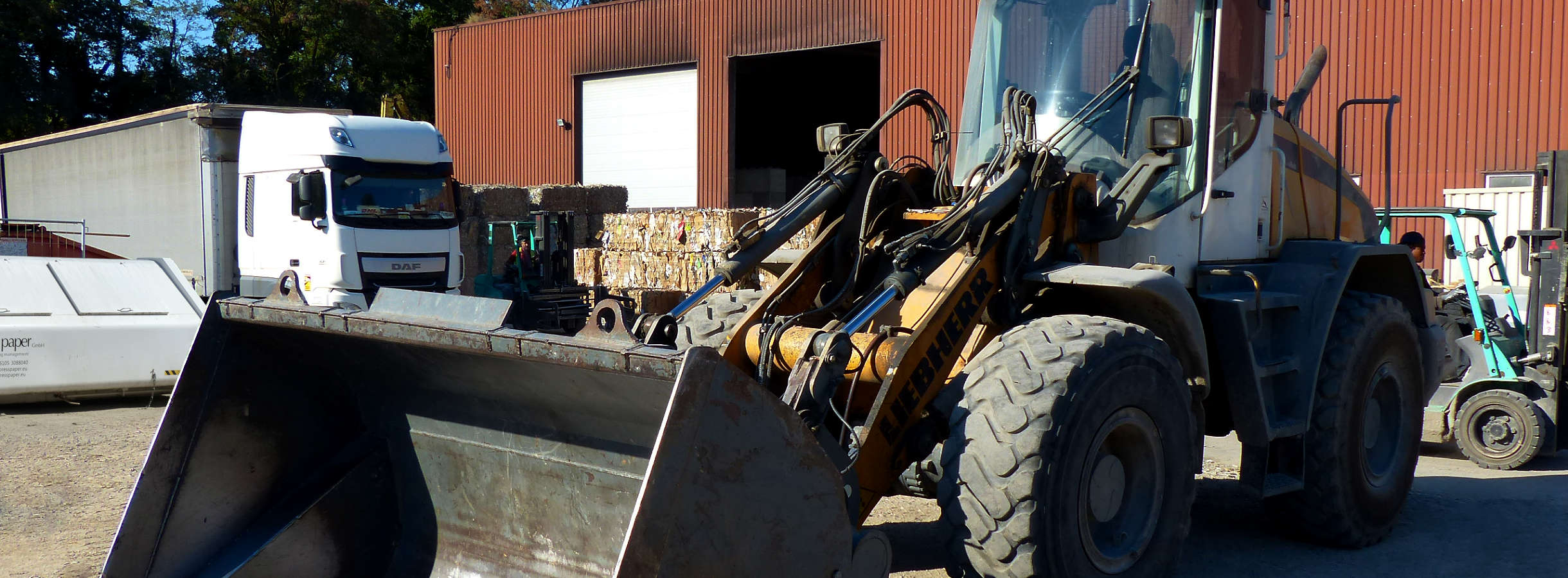 Gelände von Xpress Paper, Betriebes für Entsorgung & Recycling von Pappe, Papier und Plastik bietet viel Platz. Große Halle zum Sortieren und Pressen von Abfall. Viele gepresste Rohstoffballen. Zwei Gabelstapler laden Altpapier vom LKW. Ein Bagger ruht.