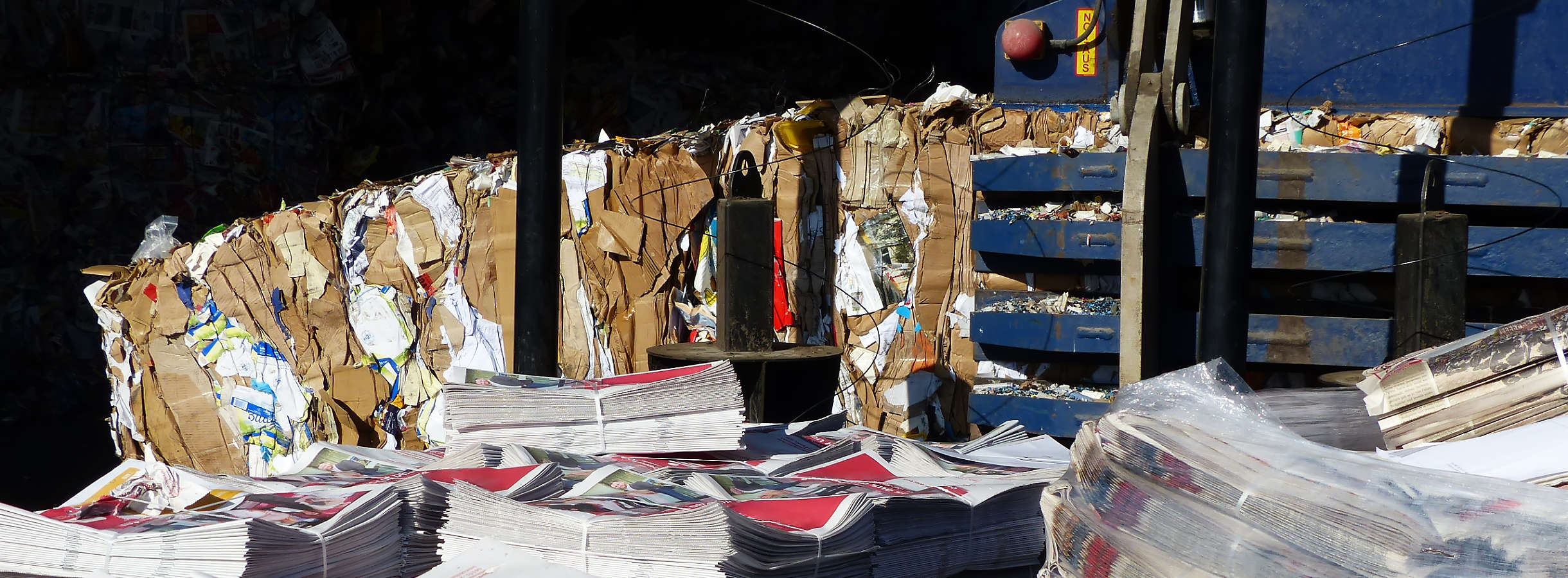 Auf Paletten gestapeltes Altpapier aus einer Werksentsorgung. Dahinter ist eine blaue Papierpresse in Aktion zu sehen. Sie presst angelieferte und sortierte Kartonage zu Karton-Ballen. Diese werden gesetzeskonform recycelt und mit Gewinn gehandelt.